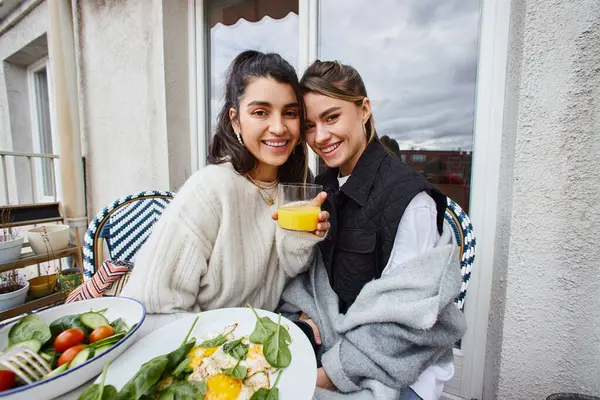 Feliz y joven pareja de lesbianas disfrutando de un desayuno saludable en el balcón, sosteniendo jugo de naranja - foto de stock