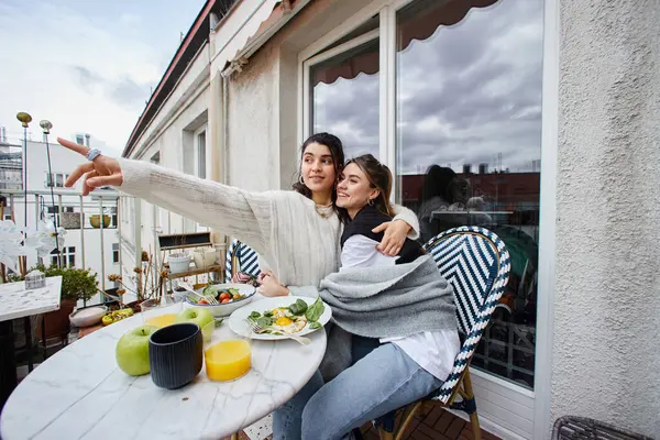 Feliz momento de joven pareja lesbiana abrazándose mientras mira hacia otro lado durante el desayuno en el balcón - foto de stock