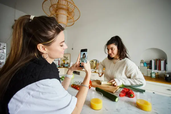 Heureux jeune lesbienne femme prise de photo sur smartphone de sa petite amie cuisine salade dans la cuisine — Photo de stock