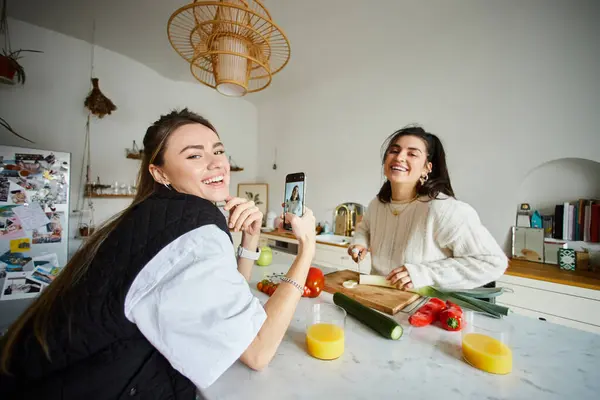 Happy young lesbian woman in 20s taking photo of her girlfriend while cooking salad in kitchen — Stock Photo