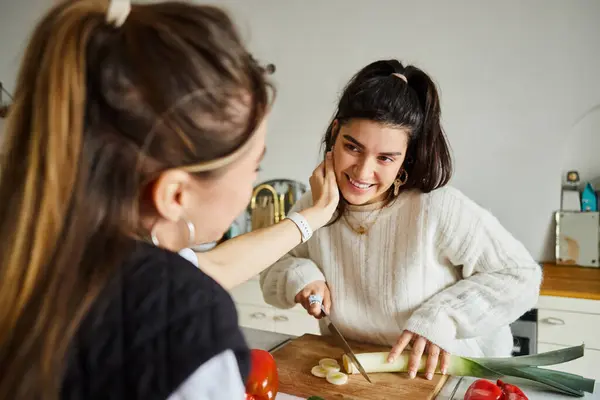 Lgbt couple, young lesbian woman adjusting hair of her happy girlfriend cutting leek in kitchen — Stock Photo