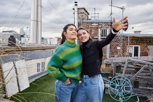 Feliz joven pareja lesbiana en traje casual de pie juntos de la azotea y mirando a la vista de la ciudad - foto de stock