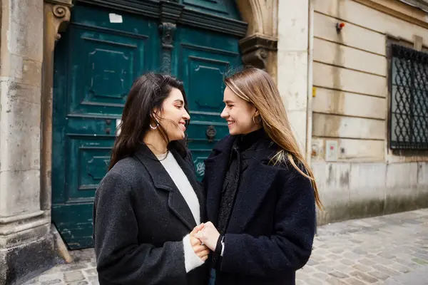 Happy lesbian couple in coats standing together and holding hands on street in European city — Stock Photo