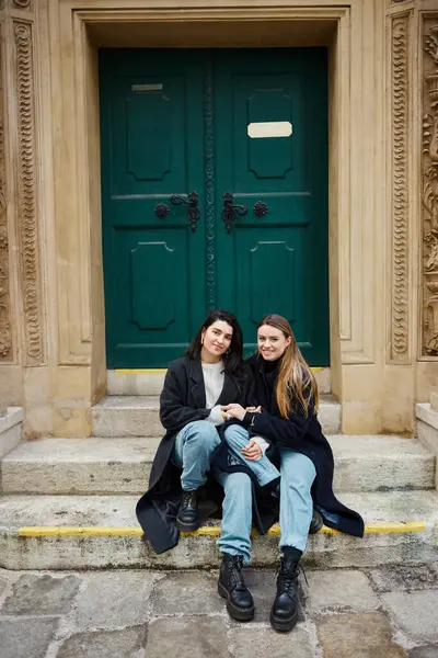 Feliz pareja lesbiana en abrigos sentados en escaleras cerca de la puerta histórica y tomados de la mano el uno del otro - foto de stock