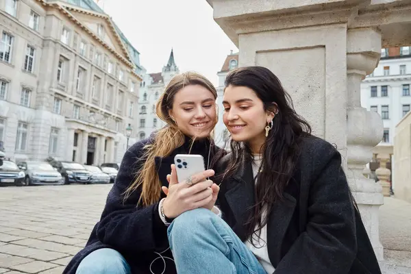 Happy and young lesbian couple using smartphone while sitting together outdoors in Vienna, Austria — Stock Photo