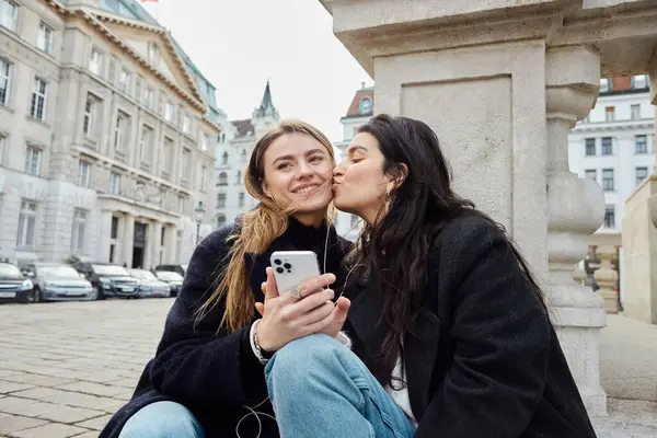 Young lesbian woman kissing cheek of happy girlfriend with smartphone while sitting outdoors — Stock Photo
