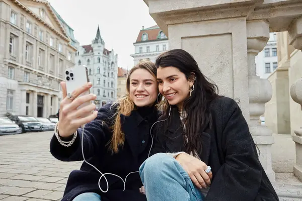 Feliz y joven pareja de lesbianas tomando selfie en el teléfono inteligente mientras se sientan juntos al aire libre en Viena - foto de stock