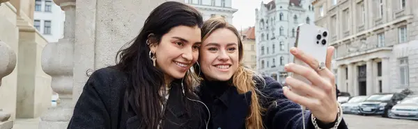Pancarta de feliz pareja de lesbianas tomando selfie en el teléfono inteligente mientras se sientan juntos al aire libre en Viena - foto de stock