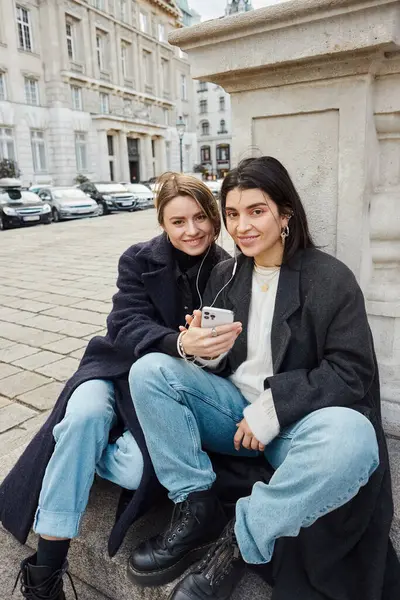 Feliz joven pareja lesbiana escuchando música en los auriculares mientras están sentados juntos al aire libre en Viena - foto de stock