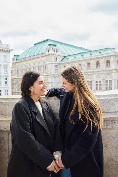 Heureux couple lesbien en vêtements de dessus souriant tout en se tenant près de l'immeuble pendant le voyage à Vienne — Photo de stock
