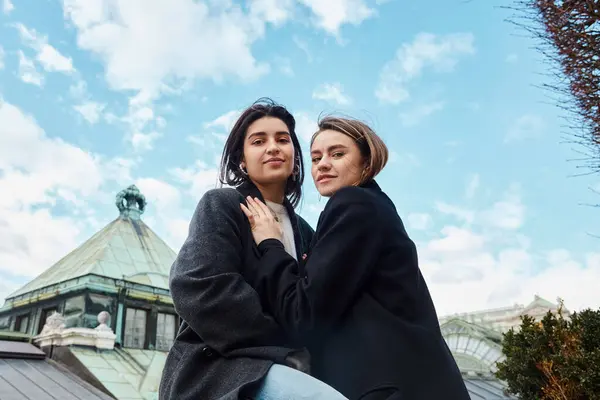 Alegre pareja lesbiana en ropa de abrigo sonriendo mientras se abrazan en Viena, mirando a la cámara - foto de stock
