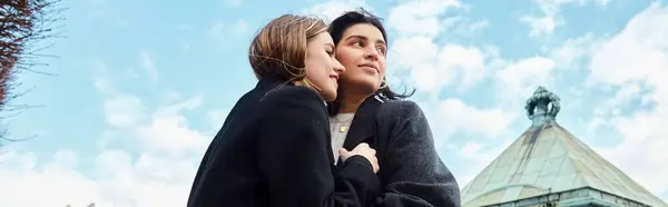 Pancarta de feliz pareja lesbiana en ropa de abrigo sonriendo mientras se abrazan cerca del edificio en Viena - foto de stock
