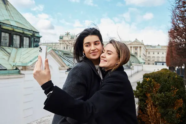 Feliz pareja de lesbianas tomando selfie en el teléfono inteligente mientras están de pie juntos en la calle en Viena - foto de stock