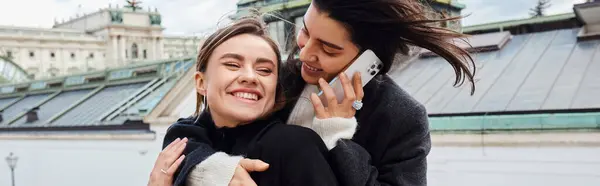 Mujer lesbiana feliz hablando en el teléfono inteligente y abrazando a su novia durante el viaje a Viena, bandera - foto de stock