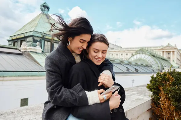 Happy lesbian woman holding smartphone and hugging her girlfriend during trip to Vienna — Stock Photo