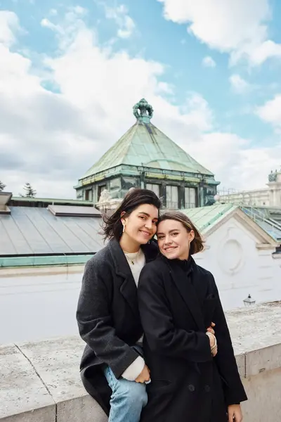 Contenida pareja de lesbianas sonriendo y abrazándose frente a una cúpula, un momento sereno en Viena - foto de stock