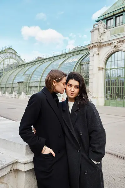 Jeune couple de lesbiennes en manteaux partageant une étreinte étroite avec Palmenhaus à Vienne sur fond — Photo de stock