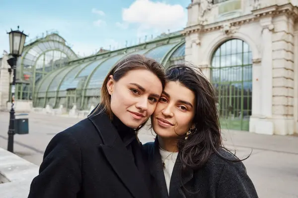 Cheerful lesbian couple in coats looking at camera near Palmenhaus in Vienna on background — Stock Photo