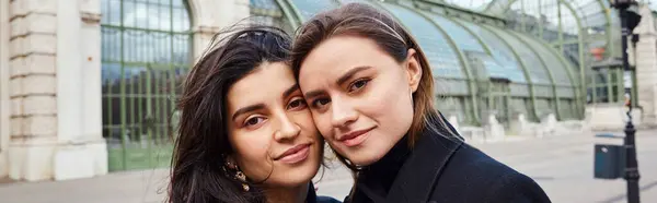 Joyeux couple de lesbiennes en manteaux regardant la caméra près de Palmenhaus à Vienne sur fond, bannière — Photo de stock