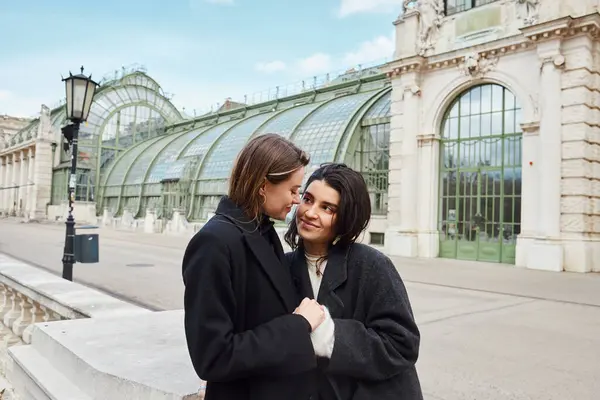 Alegre pareja lesbiana en abrigos tomados de la mano el uno del otro cerca de Palmenhaus en Viena en el fondo - foto de stock