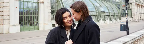 Cheerful lesbian couple in coats holding hands of each other near Palmenhaus in Vienna, banner — Stock Photo