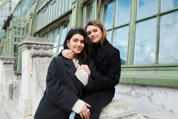 Heureux couple lesbien dans élégant noir manteaux embrasser l autre près de Palmenhaus à Vienne — Photo de stock