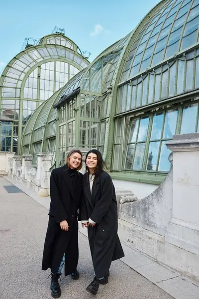 Glückliches lesbisches Paar in Mänteln Händchen haltend im Hintergrund beim Palmenhaus in Wien — Stockfoto