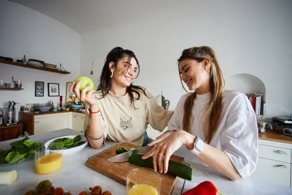 Feliz joven pareja de lesbianas sonriendo mientras hacen ensalada juntos en la cocina moderna, concepto lgbtq - foto de stock