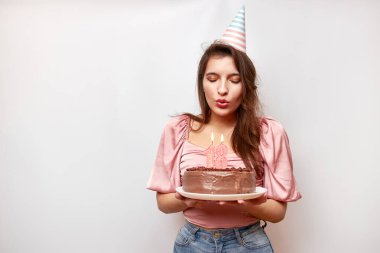 The girl is holding a festive cake and blowing out a candle in the form of the number 18. The concept of a birthday celebration.