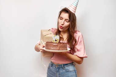 The girl is holding a festive cake and blowing out a candle in the form of the number 19. The concept of a birthday celebration.