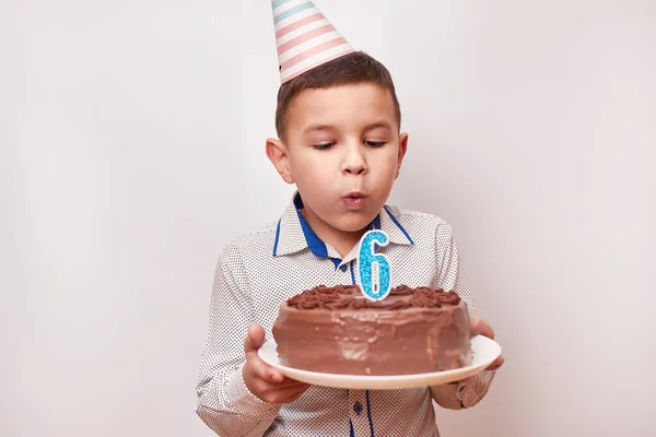 Cheerful boy holding a birthday cake blowing out a candle in the form of the number 6. Birthday celebration concept.
