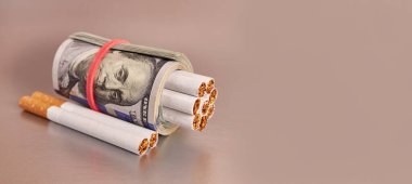 A roll of 100 dollar bills with cigarettes on a gray background. Concept of expensive cigarettes.