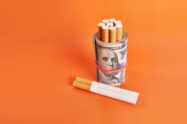 Cigarettes wrapped in 100 dollar bills on an orange background. Concept of expensive cigarettes.