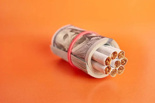 Cigarettes wrapped in 100 dollar bills on an orange background. Concept of expensive cigarettes.