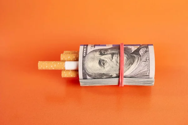 Cigarettes wrapped in 100 dollar bills on an orange background. Concept of expensive cigarettes.