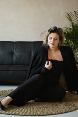 Siyah takım elbiseli bir kadın kanepenin yanındaki halıya oturur..