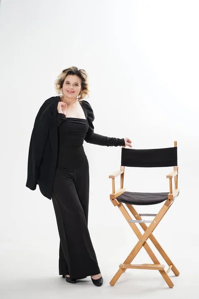 stock image A smiling woman in a black suit stands leaning on a chair on a white background.