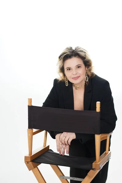 stock image A smiling woman in a black suit stands leaning on a chair on a white background.