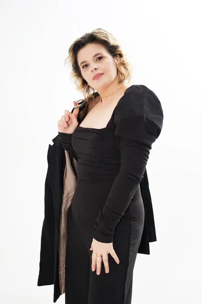 stock image A smiling woman in a black suit stands holding a jacket on her shoulders against a white background.