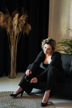 Siyah takım elbiseli gülümseyen bir kadın elinde gözlüklerle oturur..