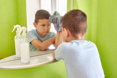 Üzgün bir çocuk aynayı bez ve deterjanla yıkıyor. Ev ödevi. Ev temizliği.