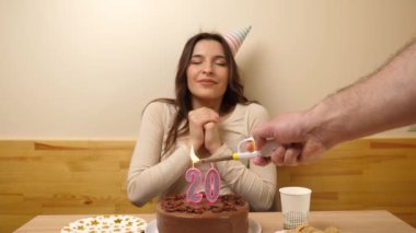 Kız elinde şenlikli bir pastayla bir masanın önünde oturuyor, 20 numara şeklinde bir mum yanıyor ve o da söndürüyor. Doğum günü kutlaması konsepti.