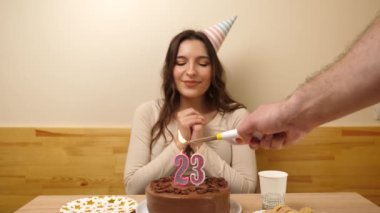 Kız elinde şenlikli bir pastayla bir masanın önünde oturuyor. 23 numara şeklinde bir mum yanıyor ve söndürüyor. Doğum günü kutlaması konsepti.