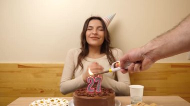Kız elinde şenlikli bir pastayla bir masanın önünde oturuyor, 27 numara şeklinde bir mum yanıyor ve söndürüyor. Doğum günü kutlaması konsepti.