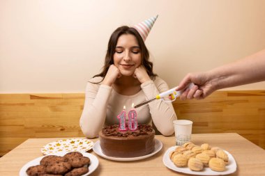 Masanın önünde elinde şenlikli bir pastayla oturan bir kız. 18 numaralı pastada bir mum yakılıyor. Doğum günü kutlaması konsepti.