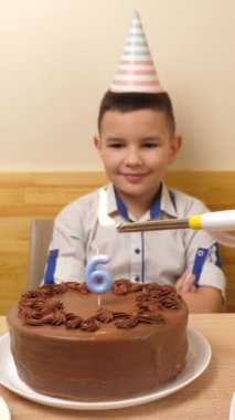Çocuk elinde şenlikli bir pastayla bir masanın önünde oturuyor, içinde 6 numaralı mumdan bir mum yanıyor ve söndürüyor. Doğum günü kutlaması kavramı. Dikey video.