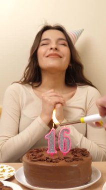 Kız elinde şenlikli bir pastayla bir masanın önünde oturuyor, içinde 16 numara şeklinde bir mum yanıyor ve söndürüyor. Doğum günü kutlaması kavramı. dikey video