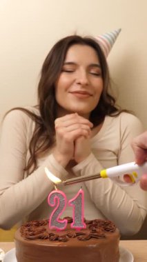 Kız elinde şenlikli bir pastayla bir masanın önünde oturuyor. 21 numara şeklinde bir mum yanıyor ve söndürüyor. Doğum günü kutlaması kavramı. dikey video