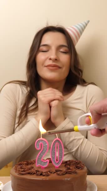 女の子はお祝いのケーキを持ってテーブルの前に座っており そこで20という数字の形をしたろうそくが燃えている 誕生日のお祝いコンセプト バーティカルビデオ — ストック動画