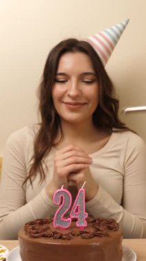 Kız elinde şenlikli bir pastayla bir masanın önünde oturuyor, içinde 24 numara şeklinde bir mum yanıyor ve söndürüyor. Doğum günü kutlaması kavramı. dikey video